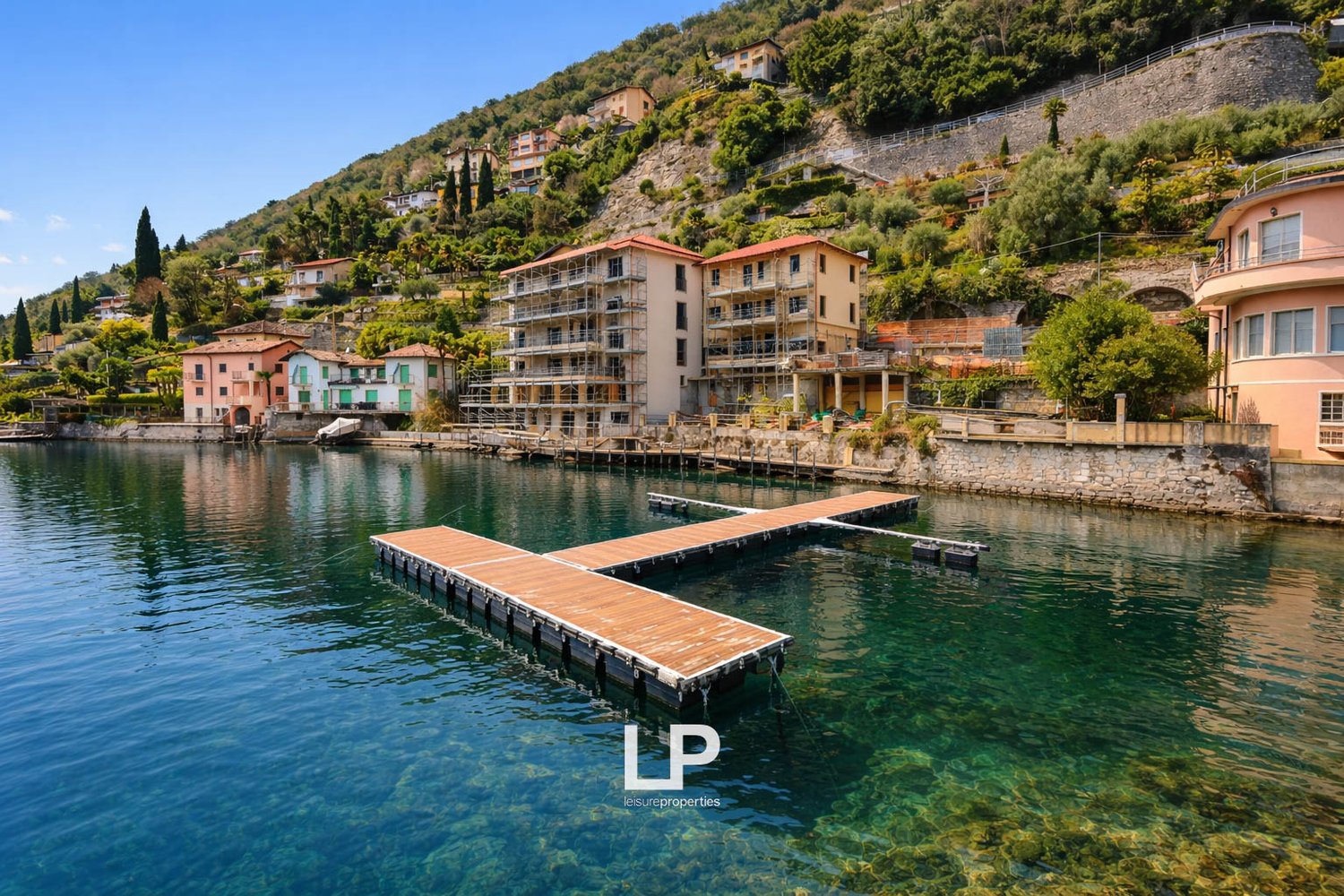 Lakeside residential complex with several buildings, a wooden dock in the foreground, surrounded by trees and a mountain in the background