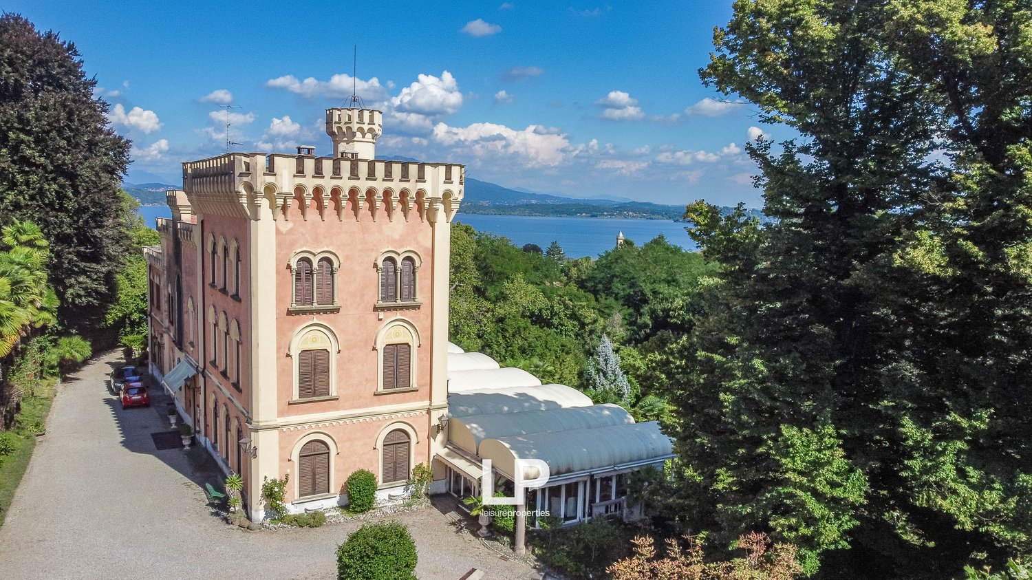 A large, pink-colored castle with turrets and arched windows, surrounded by lush greenery and overlooking a body of water in the distance.