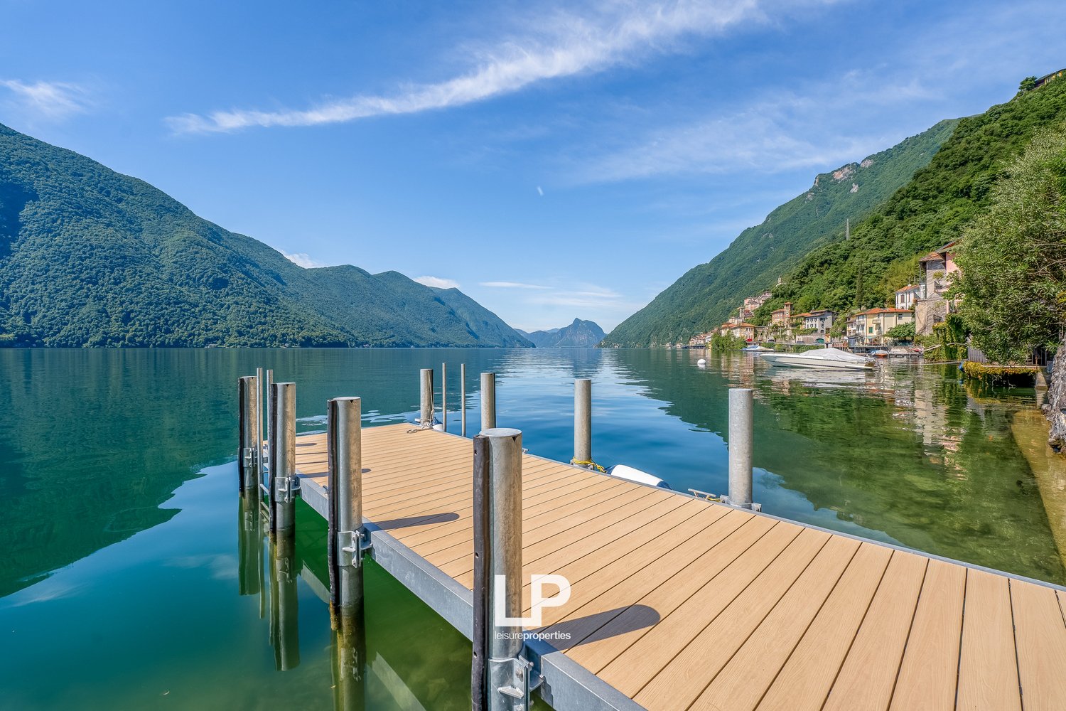Scenic view of a lake surrounded by mountains, with a wooden dock in the foreground and buildings on the shore in the distance.