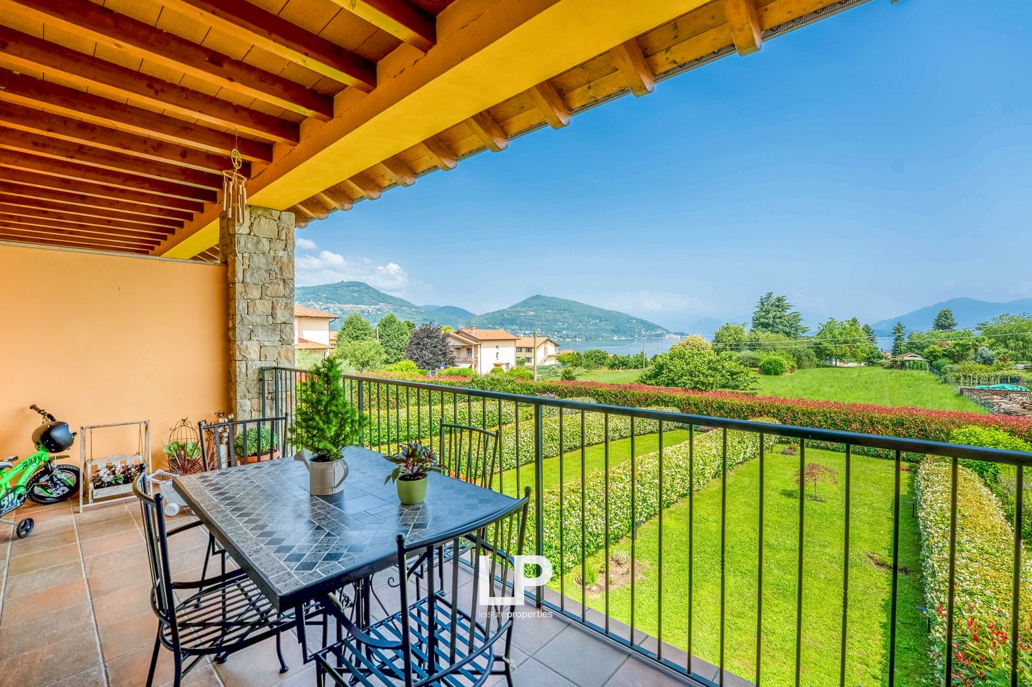 Balcony with a table and chairs, overlooking a lush green garden and scenic mountain landscape. The balcony has a wooden ceiling with exposed beams.