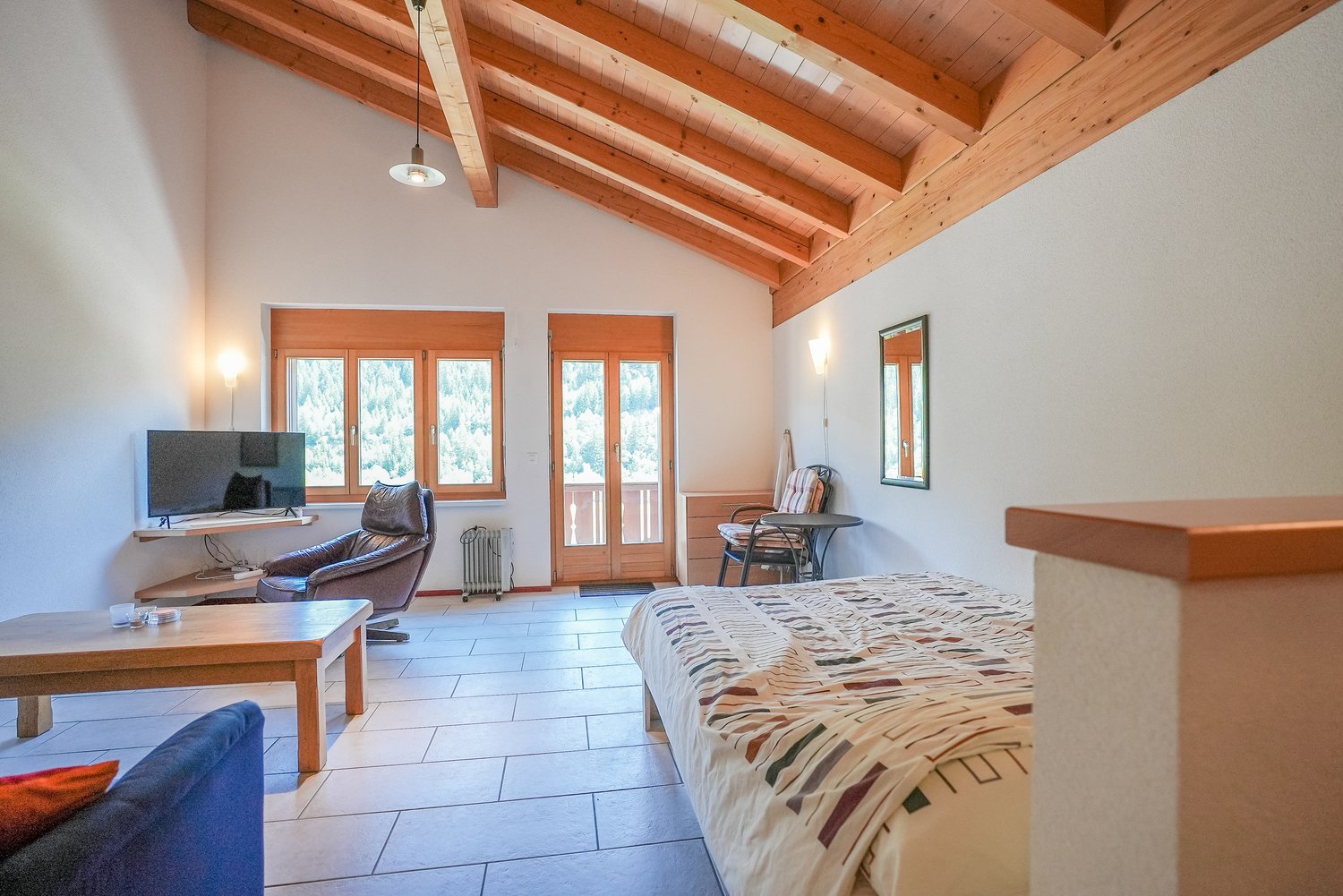 Spacious bedroom with wooden beamed ceiling, large windows providing natural light, tile flooring, leather armchair, desk, and a patterned bedspread on the bed.