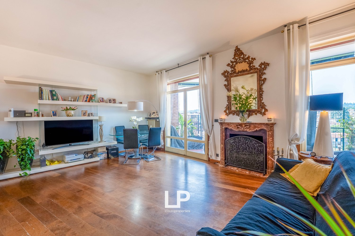 Spacious living room with hardwood floors, large windows, ornate gold-framed mirror, fireplace, TV entertainment unit, and dining area with glass table and chairs