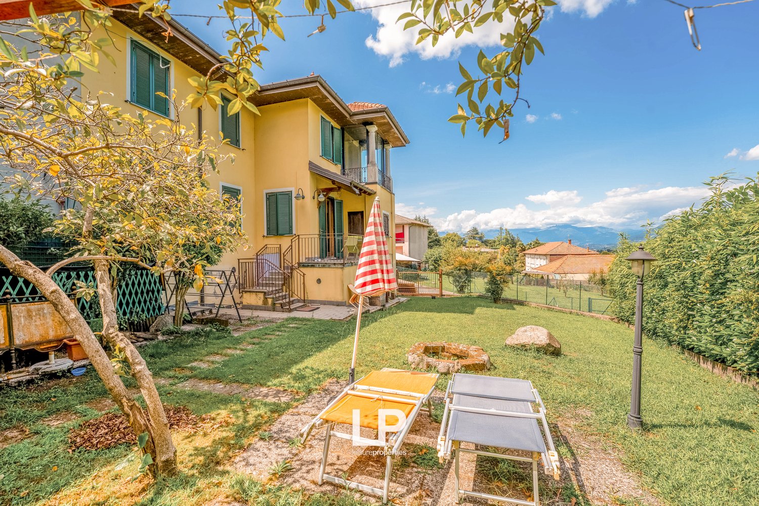 2 story house, yellow, green shutters, large backyard, deck, lounge chairs, trees, fence, mountains in the background