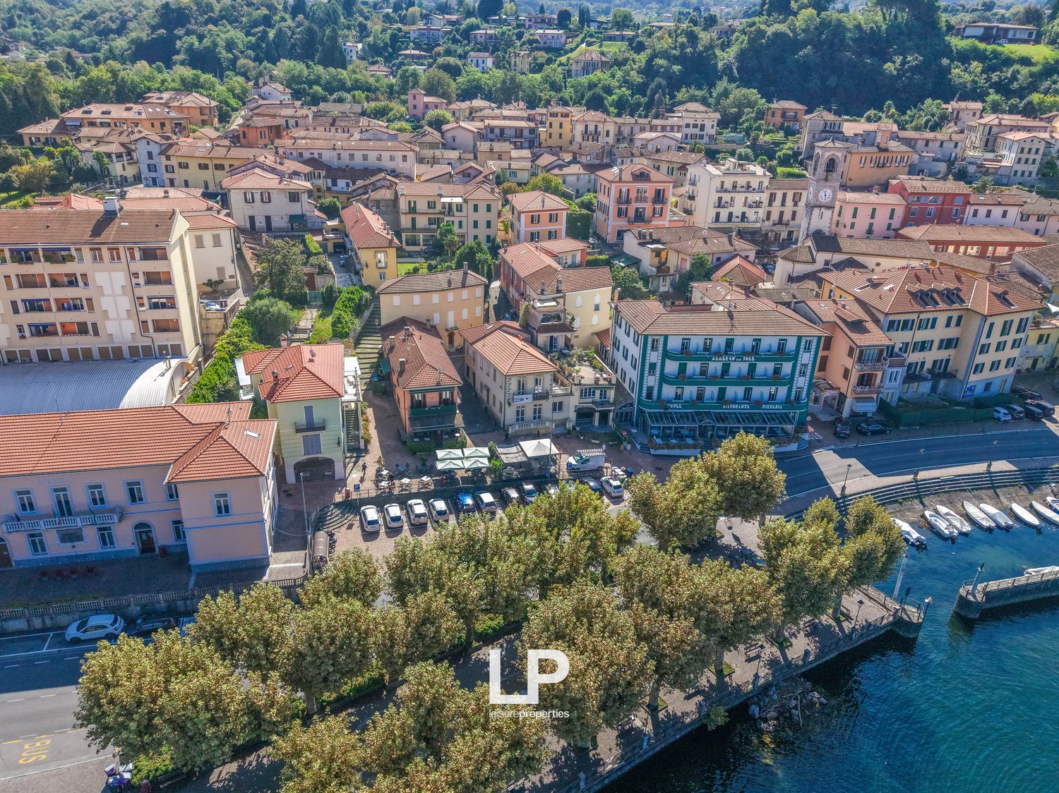 Cluster of buildings with red roofs, various sizes and styles, surrounded by trees and greenery, a winding road with parked cars, a clock tower, and a river with docked boats.