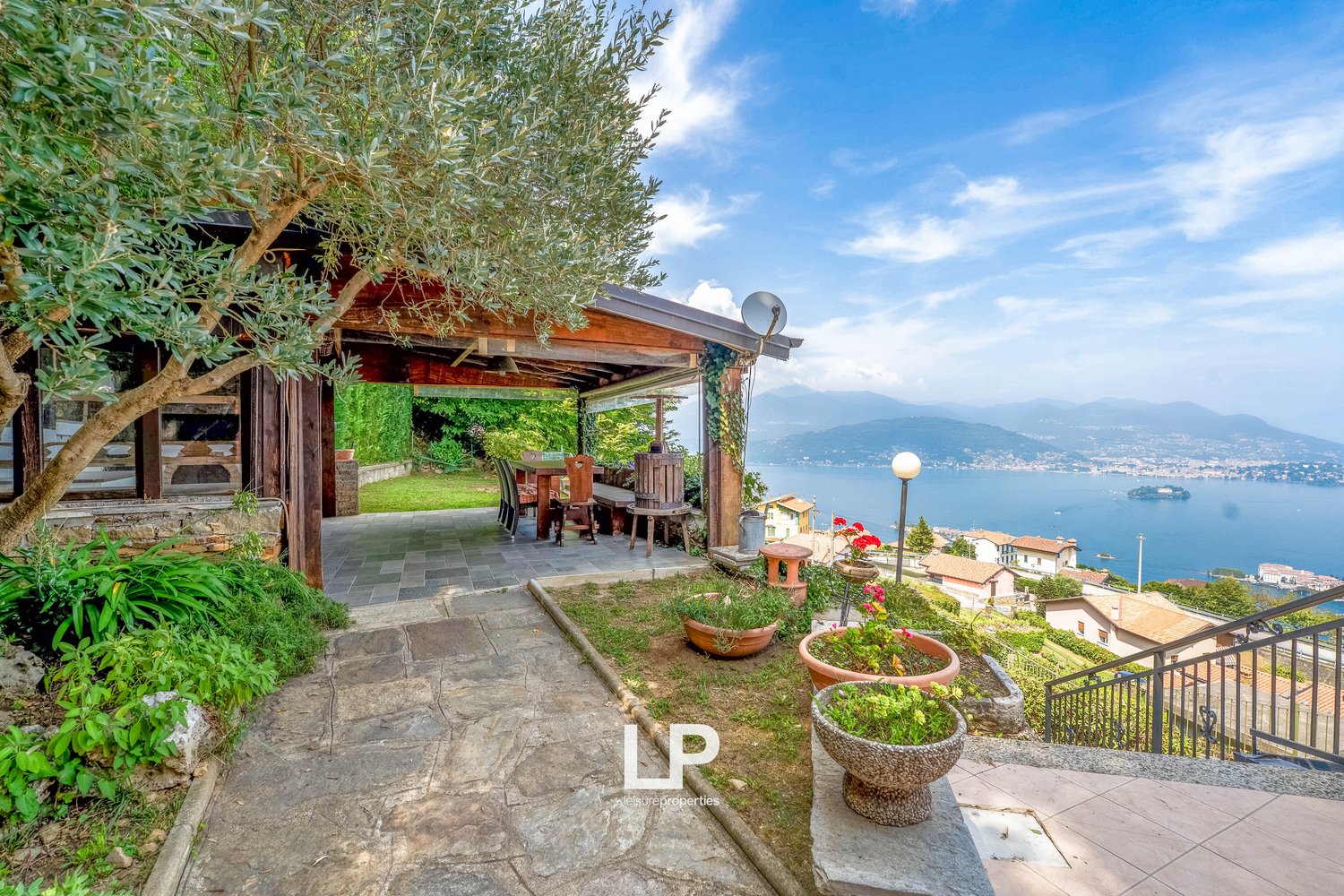 Garden with covered terrace, lake view, stairs to balcony, wooden patio table, chairs, greenery, flowers, olive tree, mountain background