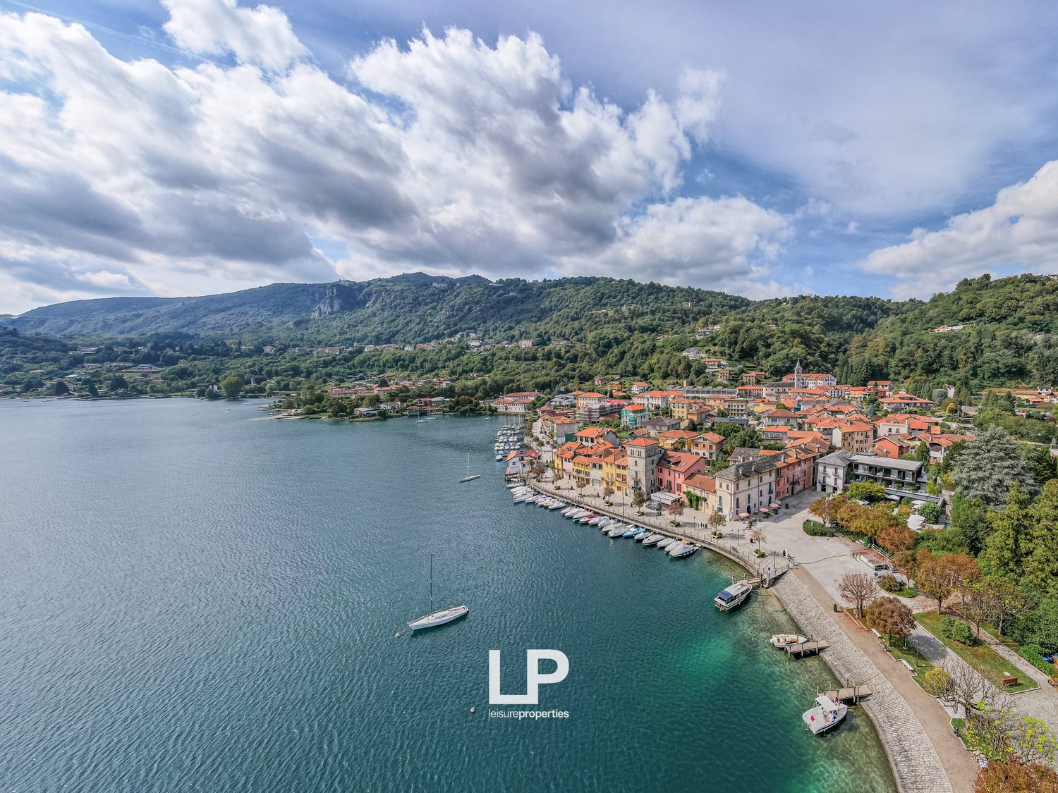 Aerial view of a coastal town with colorful houses, mountain range, boats docked, clear blue water, and trees.