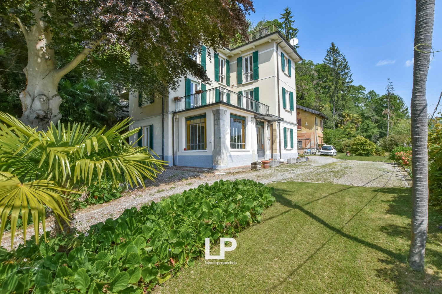 Two-story villa with a balcony, driveway, and parked car. It is surrounded by a well-maintained garden and trees. There are plants on either side of the driveway.