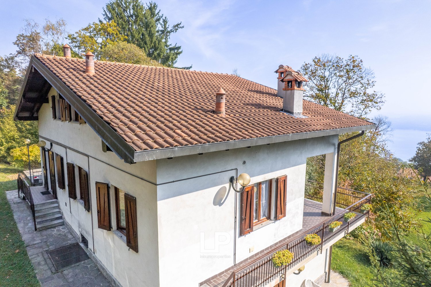 Two-story house with a brown tiled roof, wooden windows, and a tiled patio with plants. The house is raised on the ground floor and has a wooden balcony with a railing. There is a chimney on the left side of the roof.