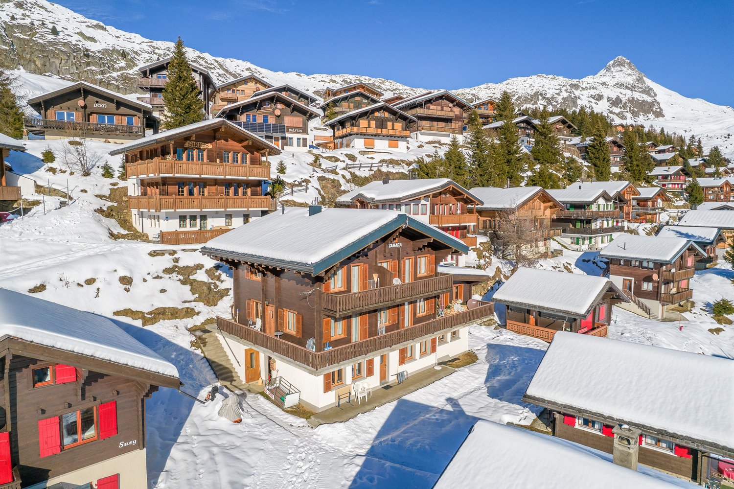 Mountain village, multiple wooden chalets, snow-covered rooftops, surrounded by trees and mountains