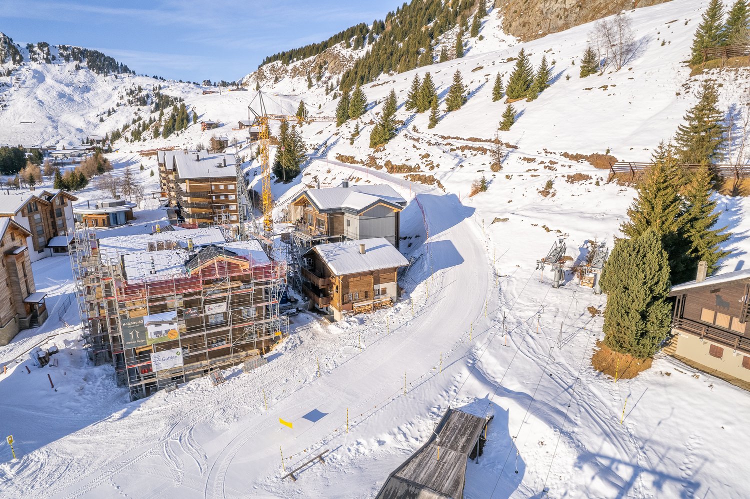 Wooden houses, snow covered mountain, ski lift in the background, trees and shrubs, a house under construction
