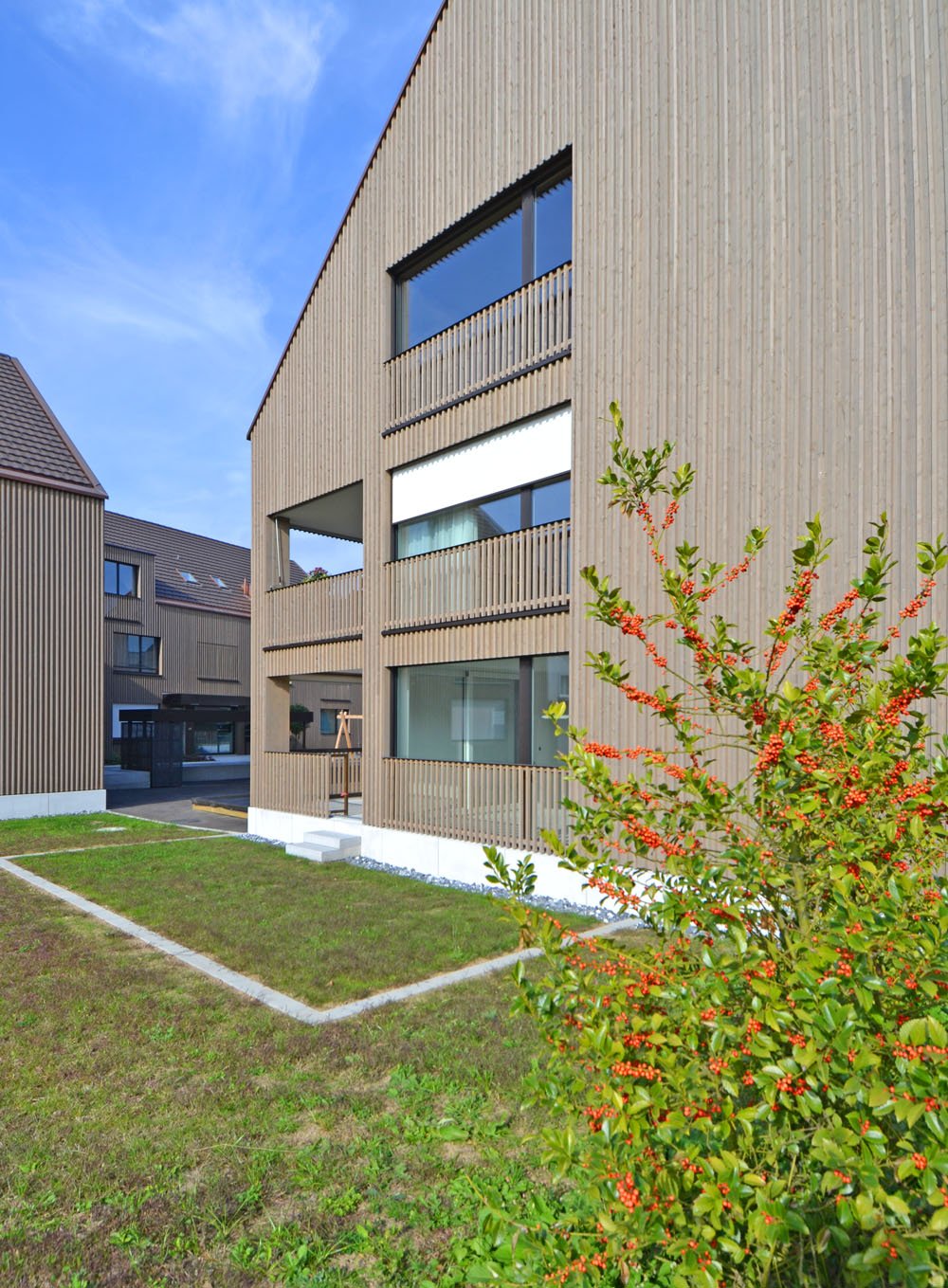 3-story modern building with wooden siding, large windows, and a balcony. The building is surrounded by a grassy area with decorative plants and shrubs.
