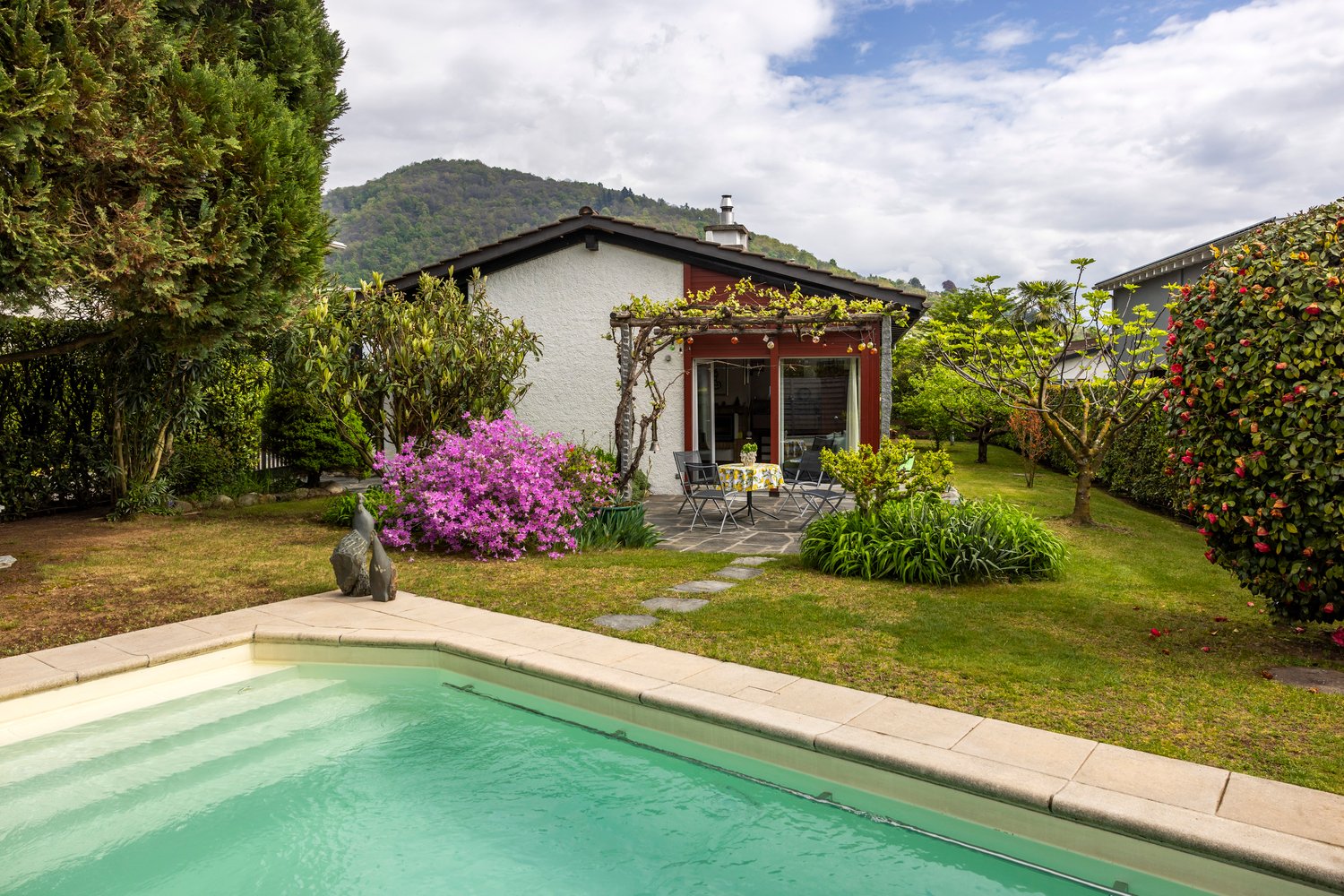 House with swimming pool, garden, patio with table and chairs, red and white exterior, mountain in the background