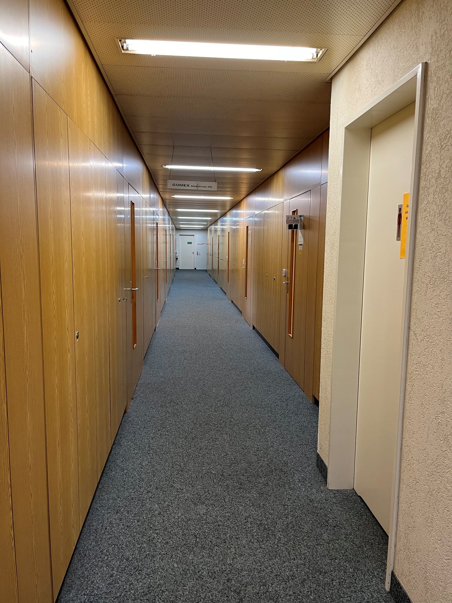long hallway with wooden lockers on both sides, gray carpet flooring, white ceilings with LED lighting, door at the end