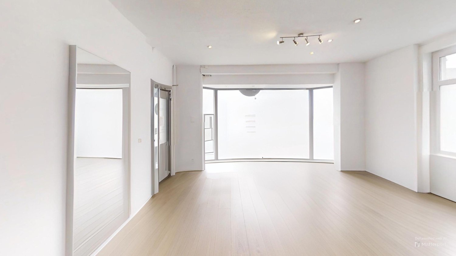 Spacious living room with large windows, hardwood floors, and track lighting. The room has a balcony or terrace visible through the windows.