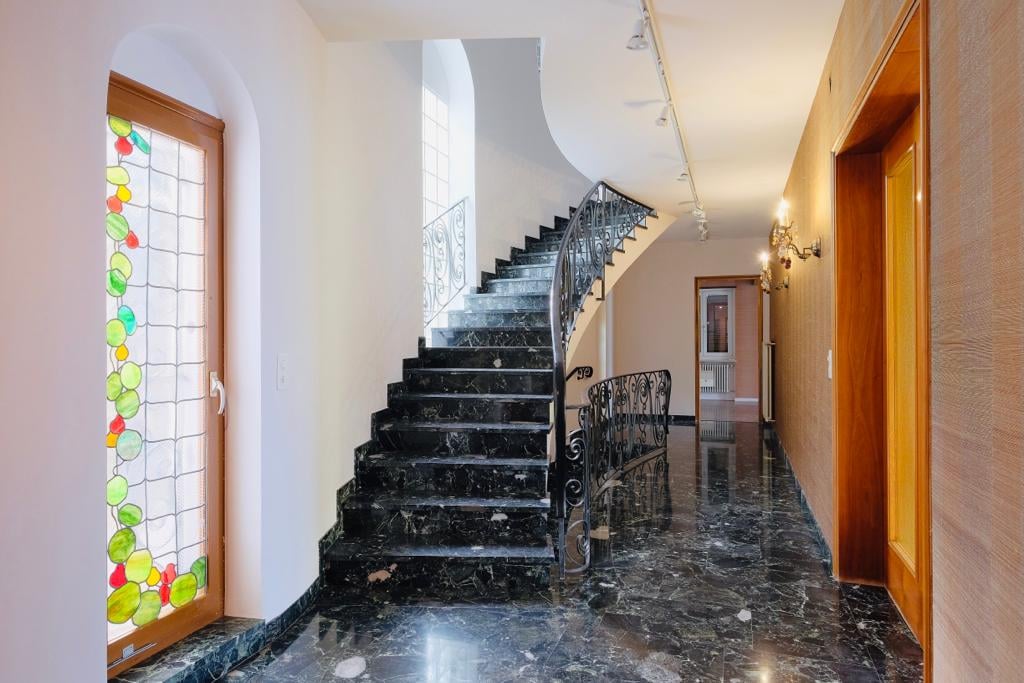 The image shows a grand staircase with black marble steps and an ornate wrought-iron railing. The walls are white, and there is a stained glass window with colorful geometric patterns. The hallway has a polished marble floor and wooden doors leading to ot