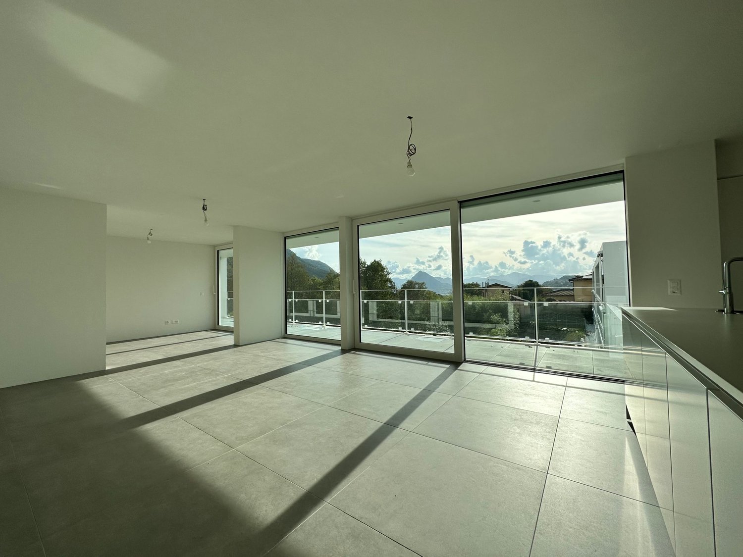 Spacious open-plan living area with large windows and sliding glass doors leading to a balcony or terrace, providing a scenic view of the surrounding mountains and landscape. The room features a minimalist design with light-colored tile flooring and white