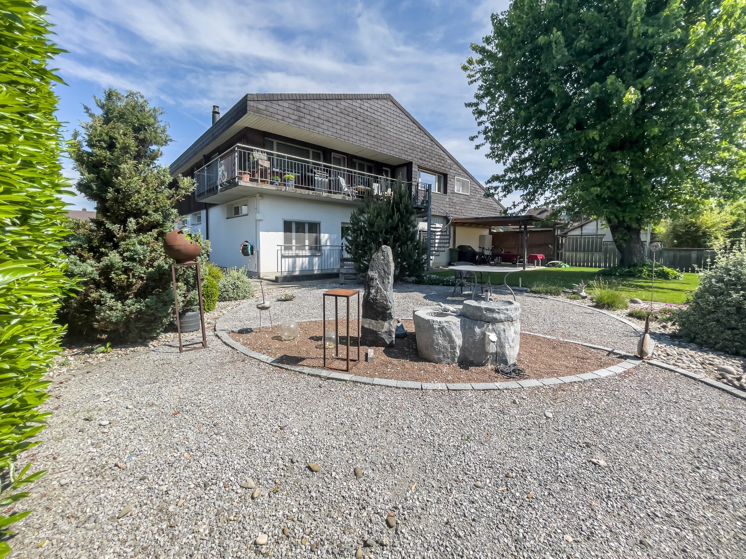 2-story house with a slanted roof, gray brick exterior, balcony, garage, and a landscaped yard with gravel, rocks, and plants
