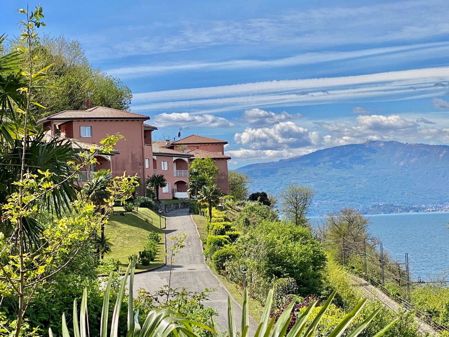 The image shows a group of multi-story residential buildings with a Mediterranean-style architecture, surrounded by lush greenery and overlooking a scenic lake or body of water. The buildings have terracotta-colored roofs and are set against a backdrop of