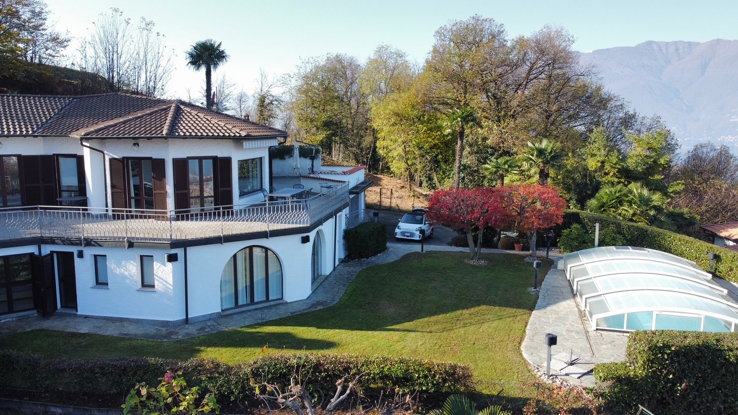Two-story white villa with tiled roof, large balcony, garage, swimming pool, surrounded by lush greenery and trees