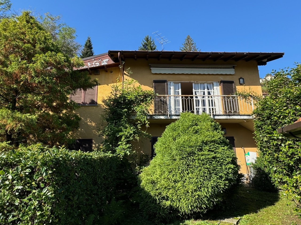 2-story yellow house with a balcony, surrounded by lush greenery and trees