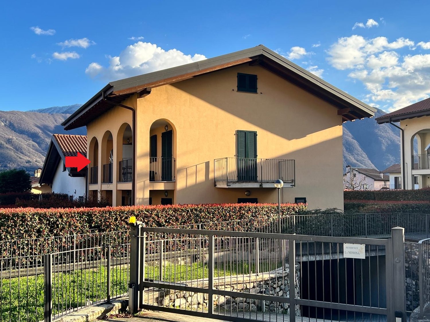2-story yellow stucco house with arched windows, balconies, and a metal roof. The house is surrounded by a metal fence and landscaping with flowers and shrubs.
