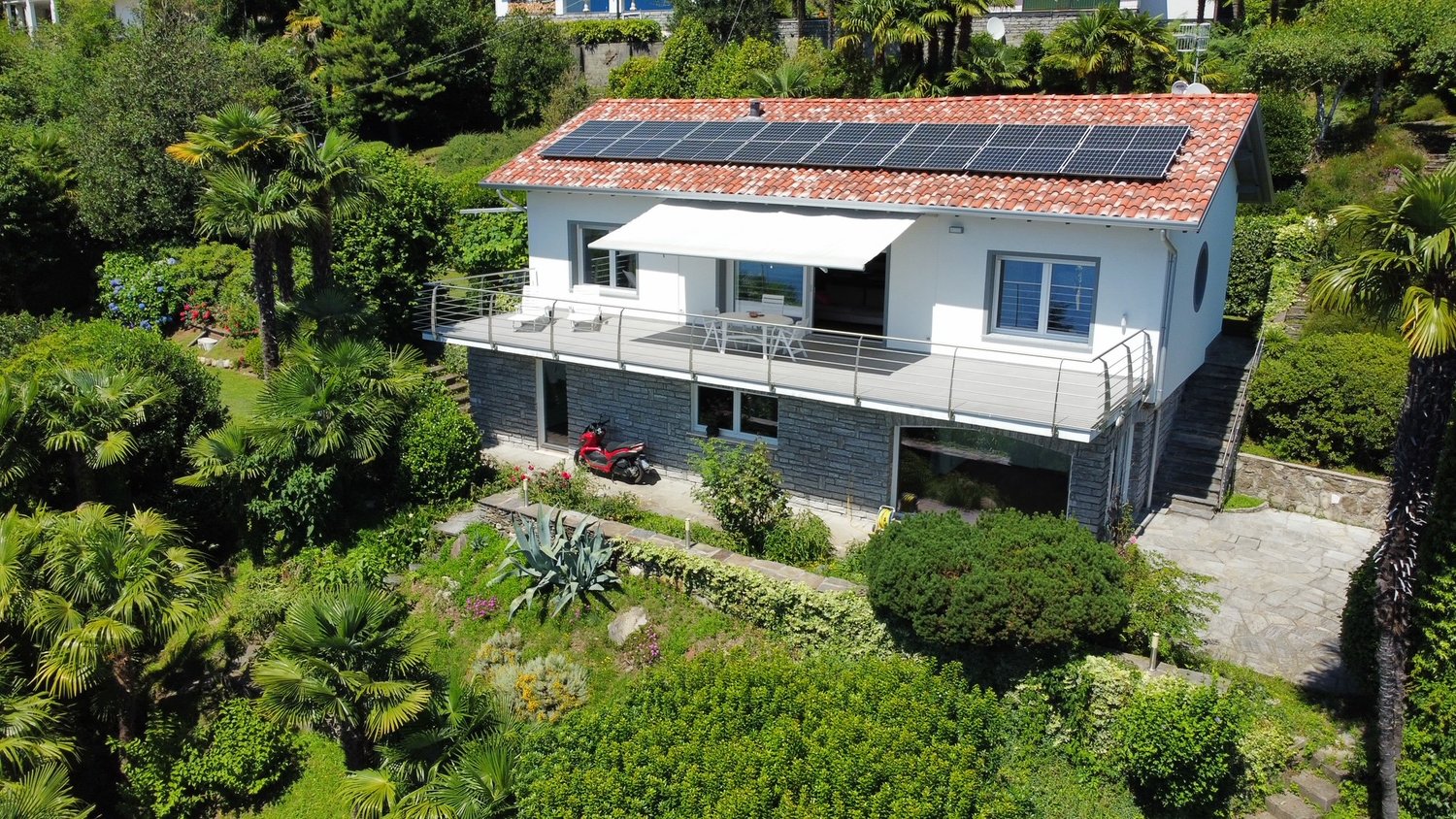 2-story modern house with a tiled roof, solar panels installed on the roof, a balcony or terrace, and surrounded by lush greenery and palm trees.