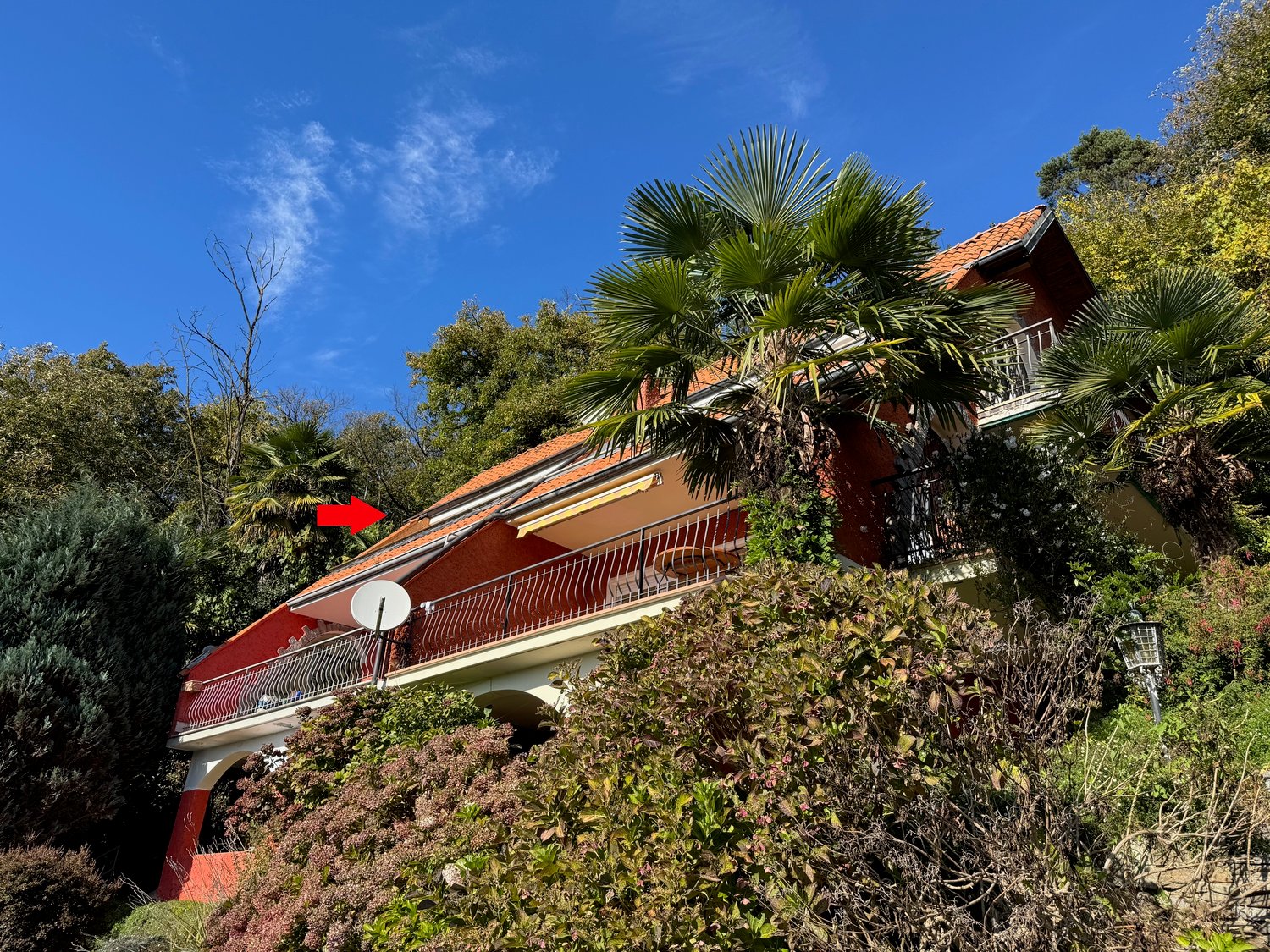 Red and white house, large balcony, red roof, balcony railing, surrounded by greenery and trees