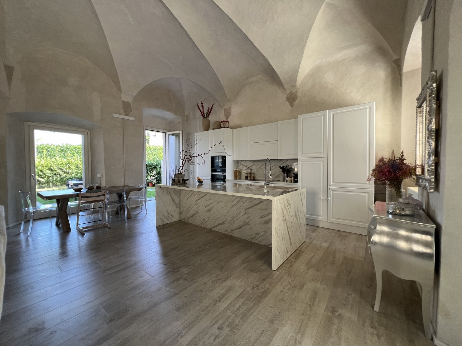 white modern kitchen with marble countertop, central island with sink, dining table with chairs, window with view to garden