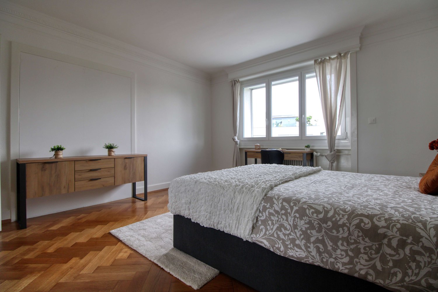 Bedroom with wooden floor, white walls, bed, wooden side table, desk, chair, window with curtains, rug, and potted plants.