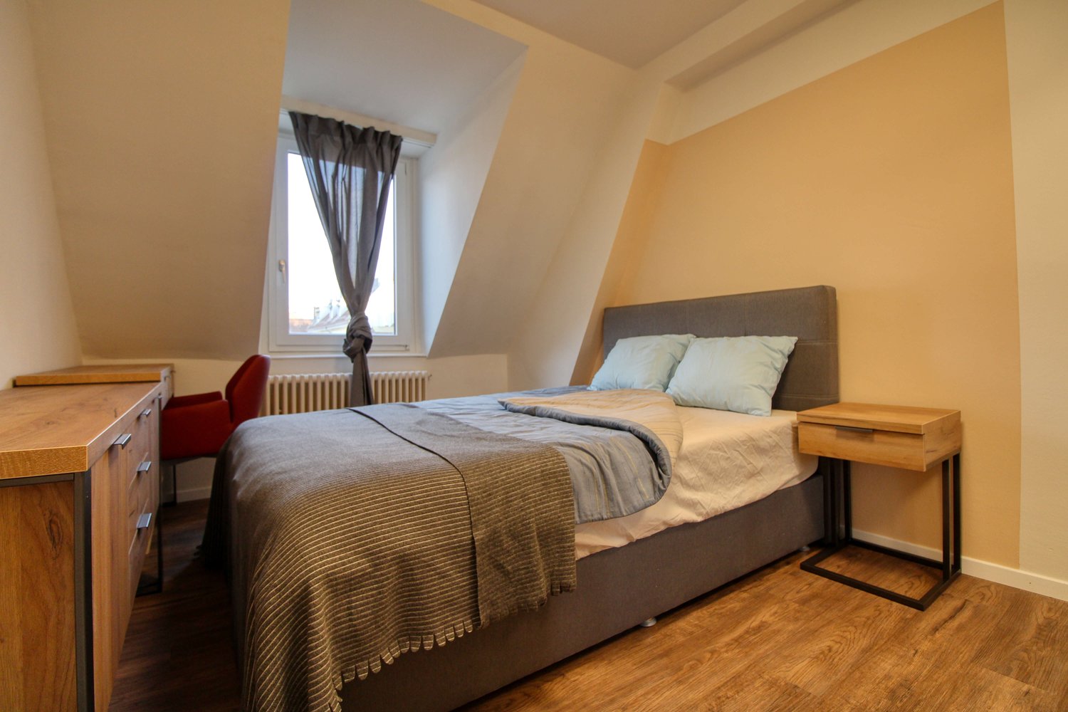 Bedroom with a bed, two nightstands, a desk, a chair, a window with curtains, and a radiator. The bed has a duvet and two pillows, and the desk has drawers.