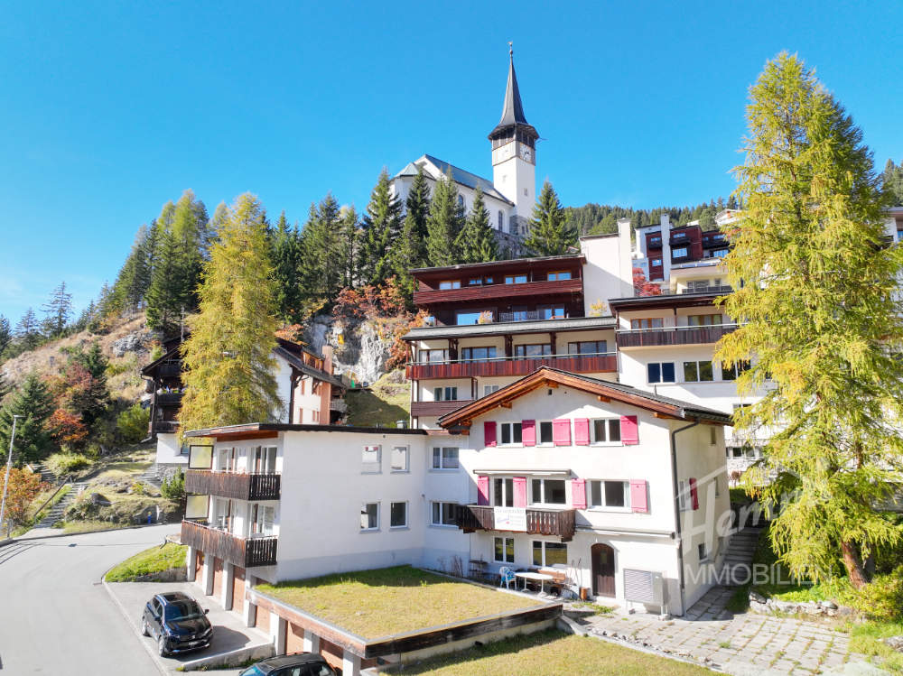 Mountainside building with multiple balconies, green roof, church in the background, surrounded by trees