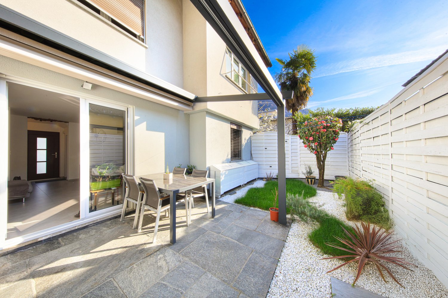 Two-story house with a patio area, outdoor dining set, and landscaped garden with palm trees and other plants. The house has large windows and a sliding glass door leading to the patio. The patio is paved with stone tiles and has a gravel area with potted