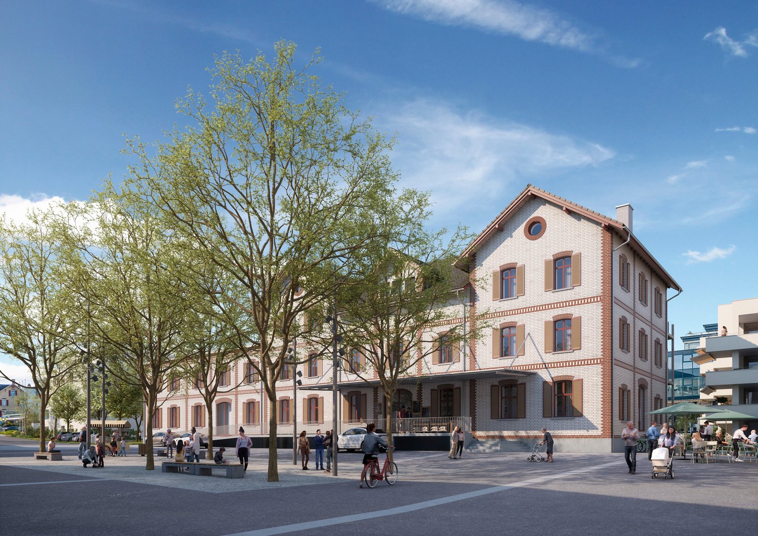 A large, multi-story brick building with arched windows and a red tile roof. The building is surrounded by trees and is situated in a public square with people walking, sitting, and riding bicycles.