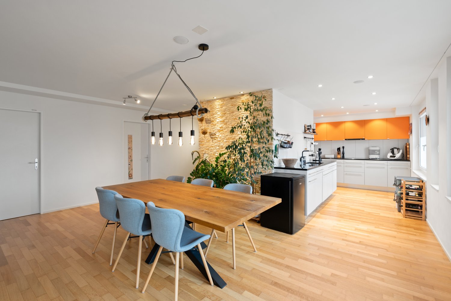 dining table, wooden chairs, kitchen island with cooking area, open shelves, modern pendant light, orange cupboards, wooden flooring