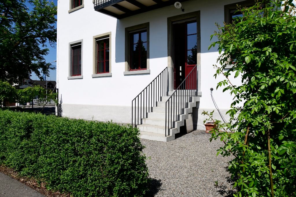 white house, red door, concrete steps, gravel driveway, hedges, trees