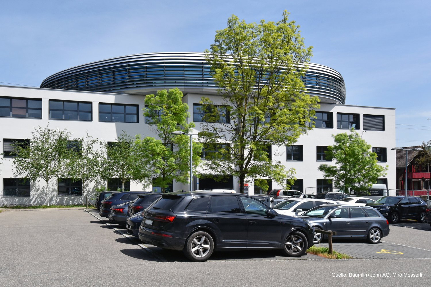 A modern, multi-story office building with a curved, domed roof structure. The building has large windows and is surrounded by trees. There is a parking lot in front of the building with several cars parked.