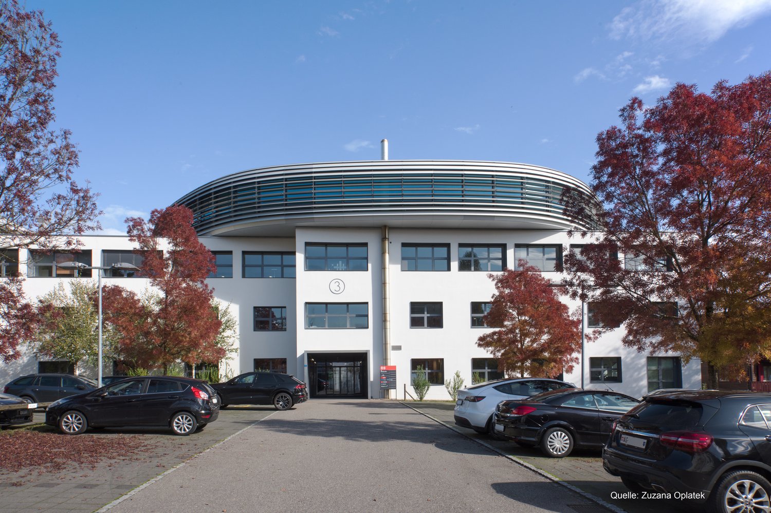 A modern, multi-story office building with a curved, domed roof structure. The building has large windows and is surrounded by trees with red autumn foliage. There is a parking lot in front of the building with several cars parked.
