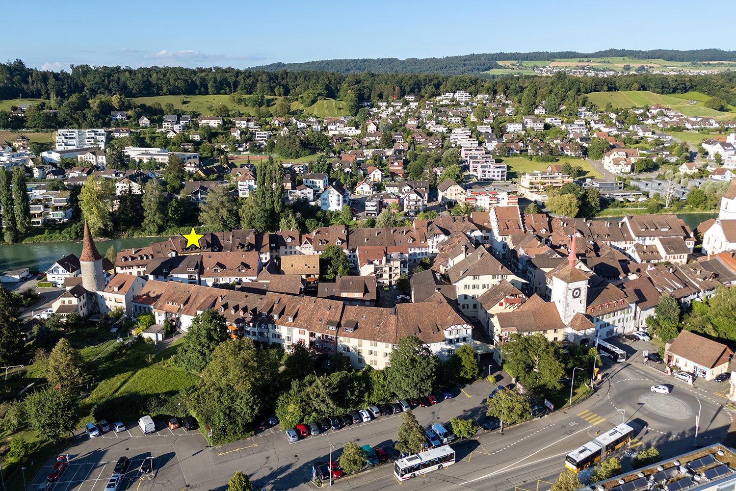 Aerial view of a large building complex with multiple buildings, brown roofs, and a yellow star indicating a specific building. The complex is surrounded by trees, grass, and a river. There are also several cars parked in the parking lot.