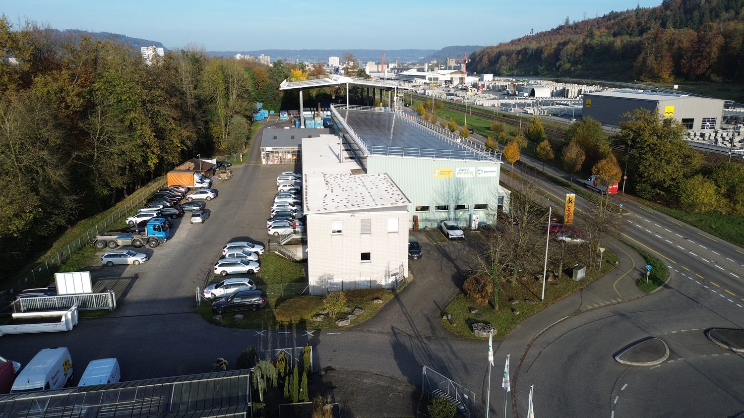 An industrial facility with a large parking area with multiple vehicles parked. A warehouse with a solar panel roof is situated on the property. Several buildings and trees are in the distance.