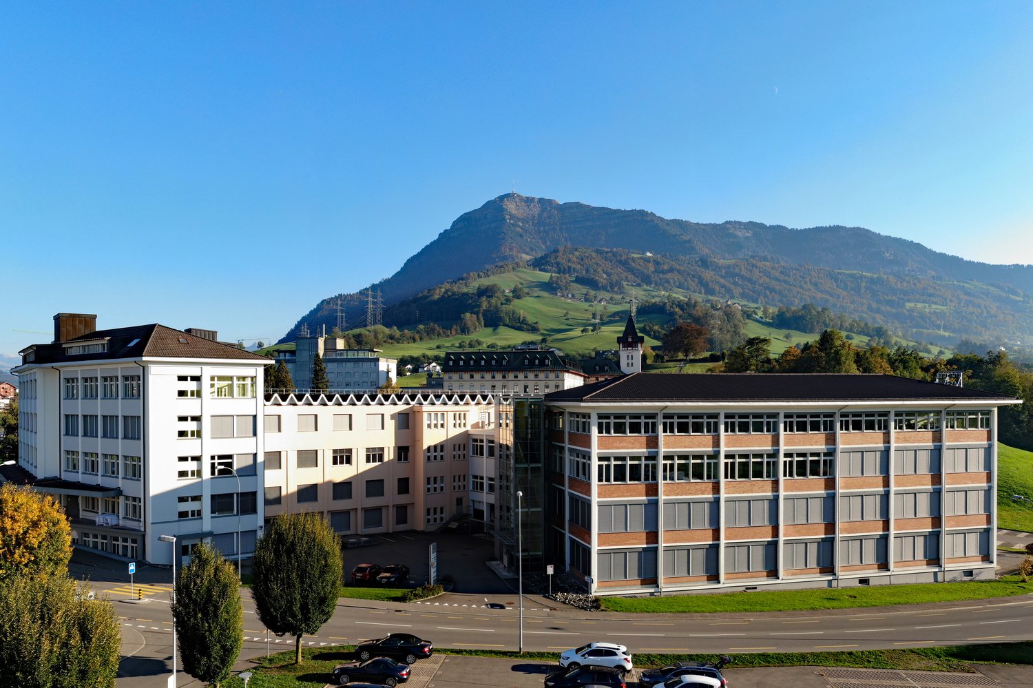 2 building structures, multiple cars parked, green grass and trees, view of a mountain