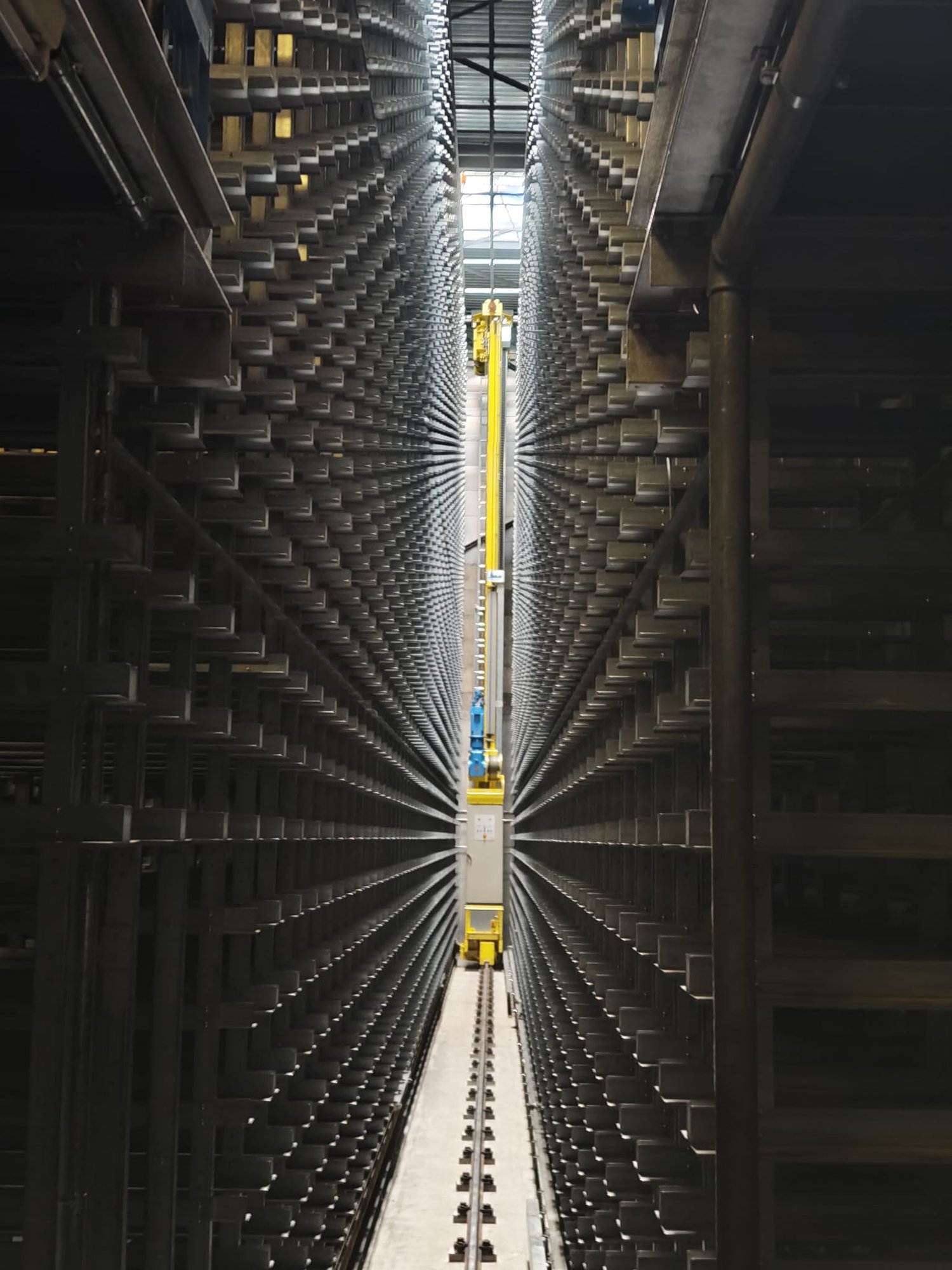 Industrial facility with large metal shelving units, yellow automated retrieval system, and a central aisle