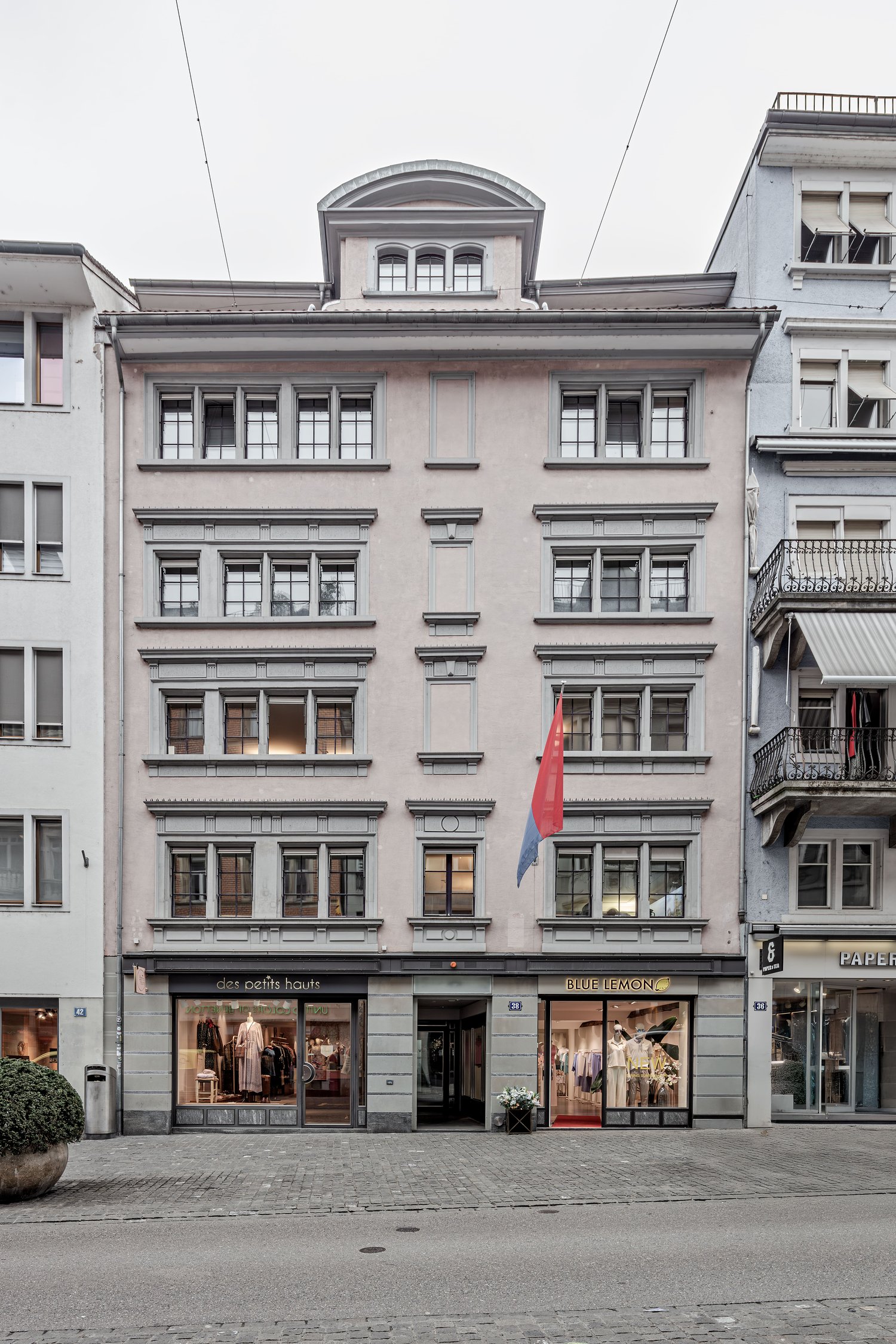 A multi-story building with a classical architectural style, featuring arched windows, decorative moldings, and a central pediment. The building has several retail stores on the ground floor, including 'Des petits hauts' and 'Blue Lemon'. A Serbian flag i