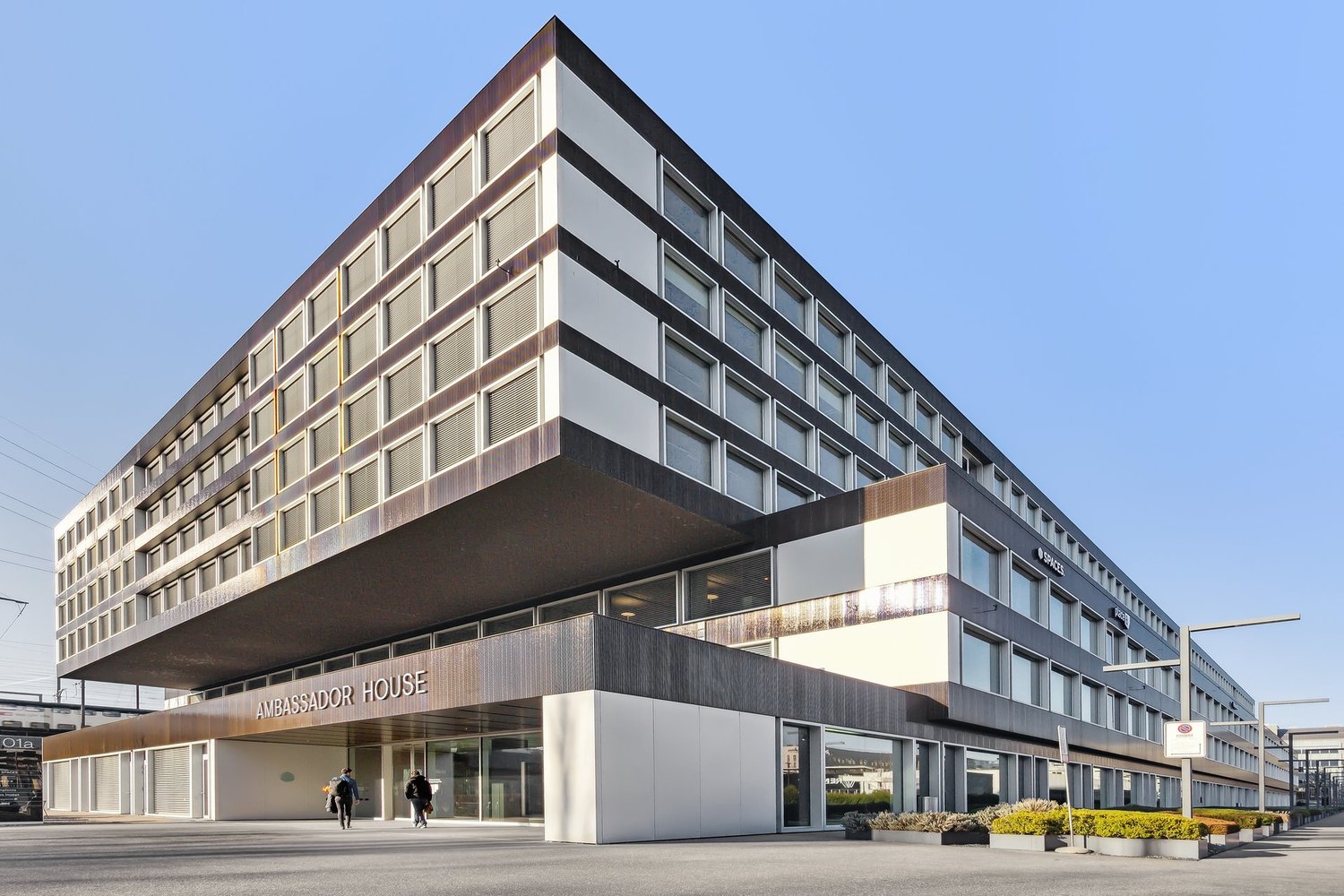 A modern, multi-story office building with a sleek, angular design. The building has a large, prominent sign that reads 'Ambassador House' on the front facade. The exterior is composed of a mix of glass, metal, and concrete materials, creating a contempor