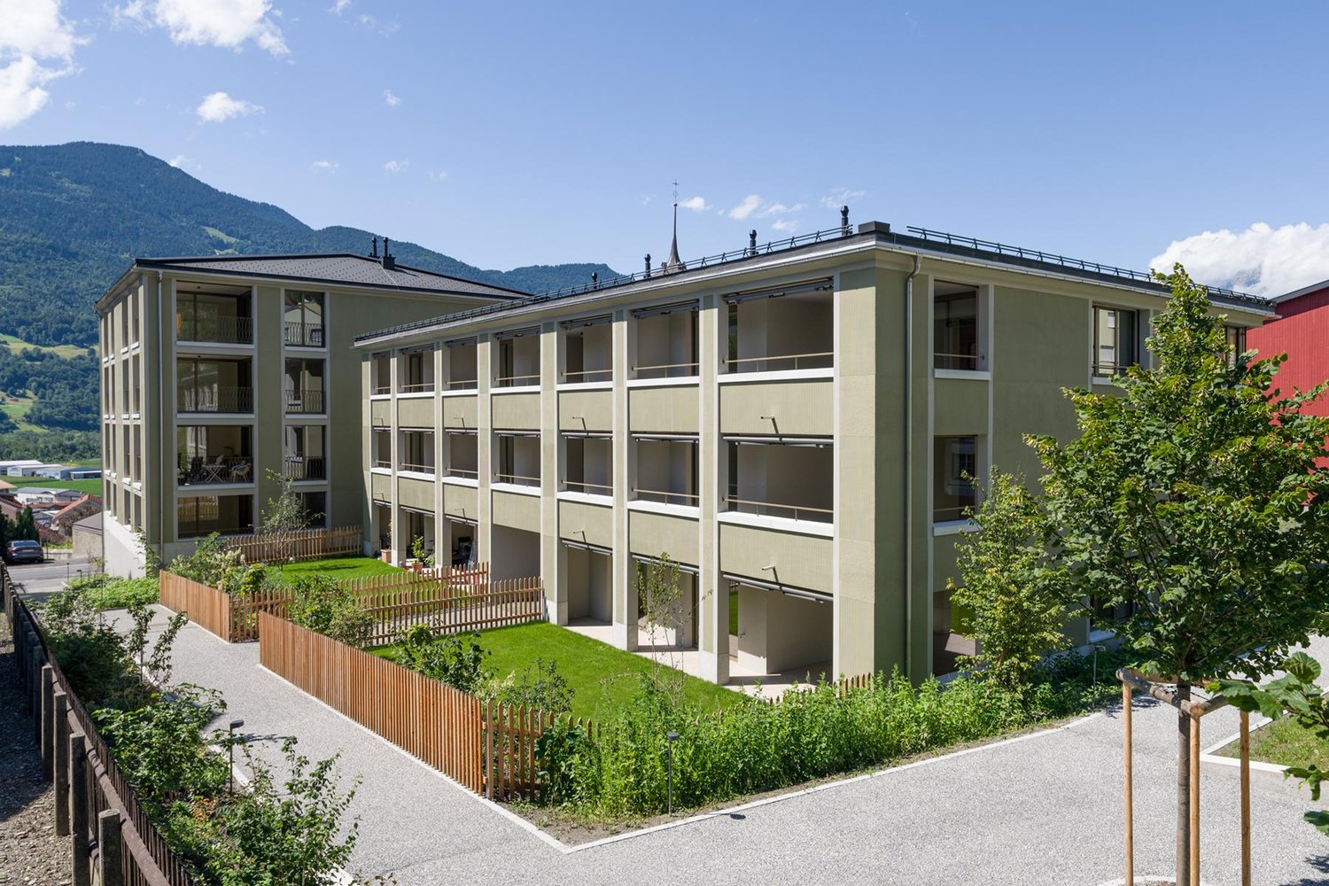Multi-story apartment building with balconies, surrounded by landscaping and fencing, located in a mountainous area with a blue sky and clouds in the background.