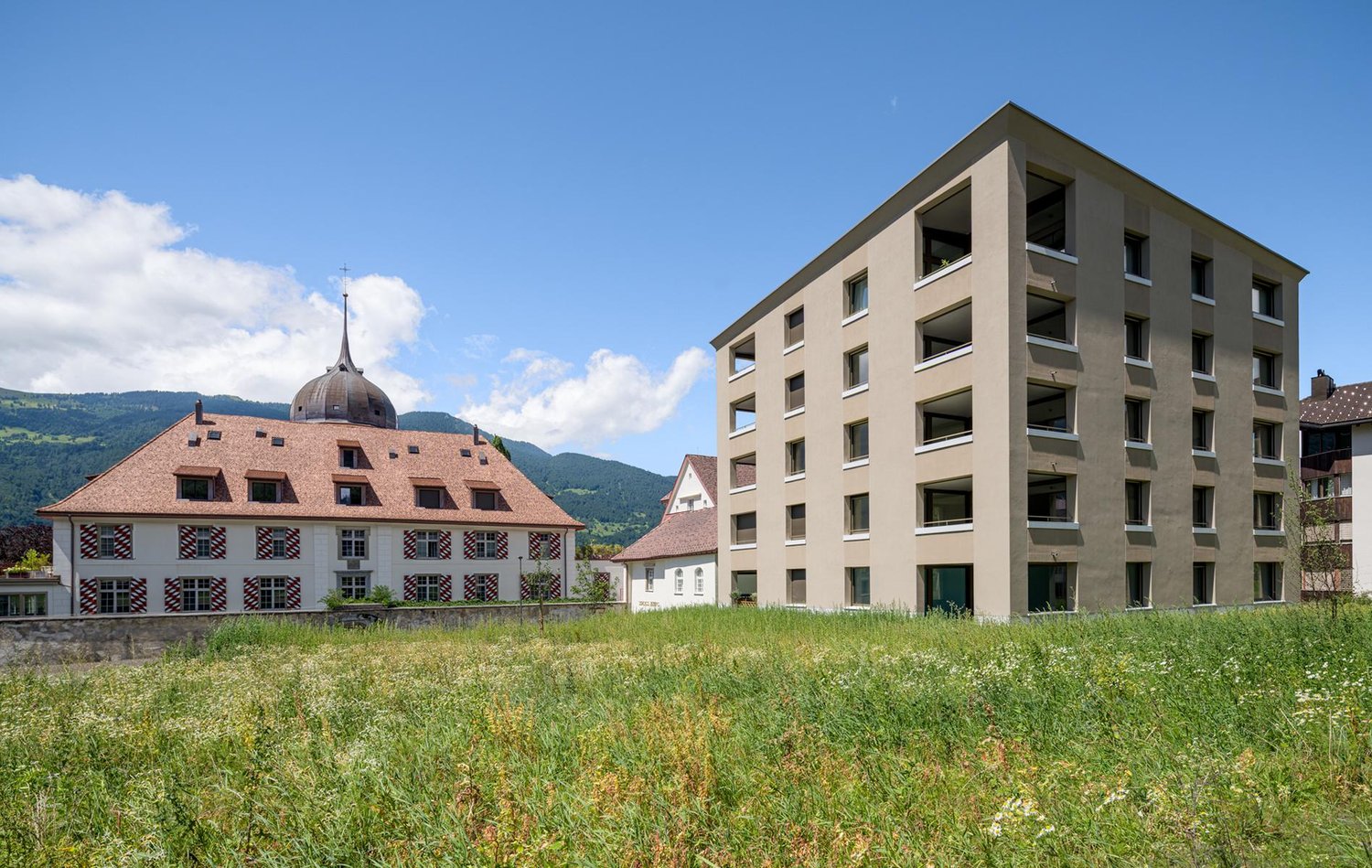 The image shows a modern multi-story apartment building with a traditional historic building in the background. The apartment building has several balconies and is surrounded by a grassy field with mountains in the distance.
