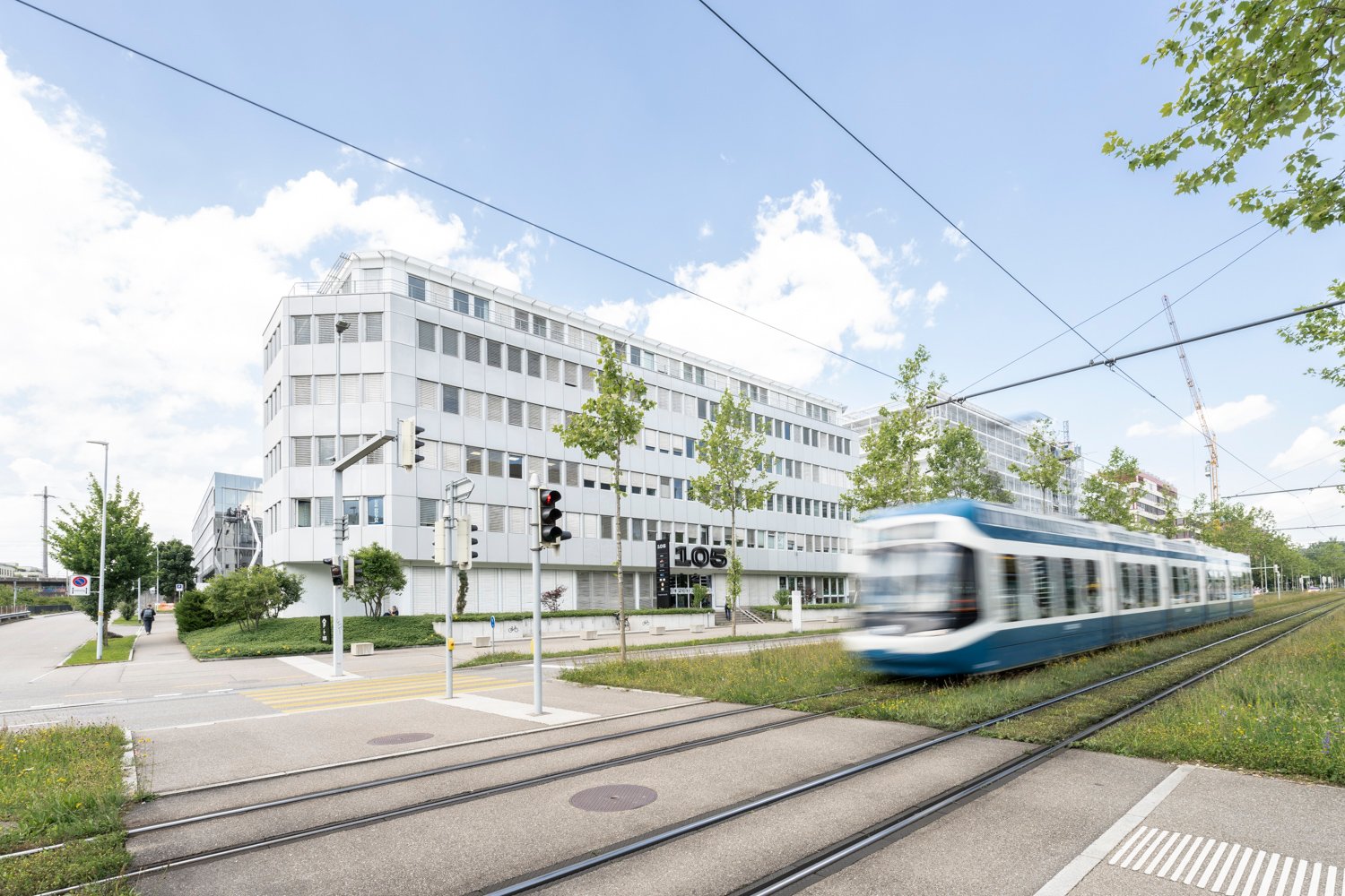 A multi-story commercial building with a modern, white facade. There are trees and a tram track in the foreground, and a tram is visible passing by.