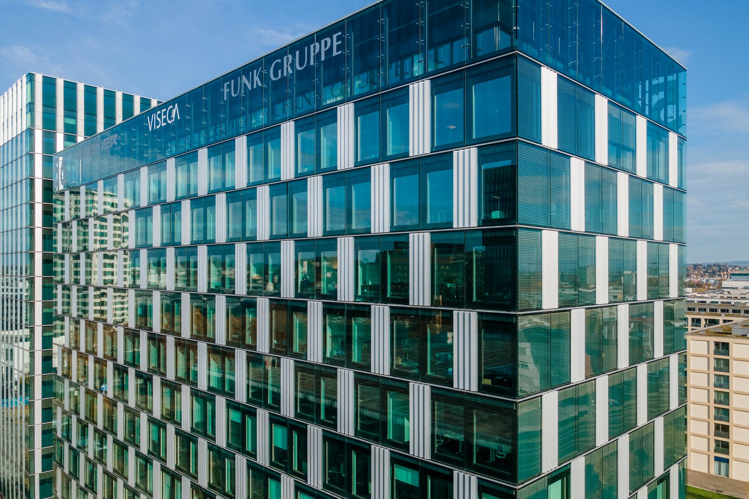 Large modern office building with glass facade, Funk Gruppe logo visible, multiple floors, parking spaces visible in front of the building