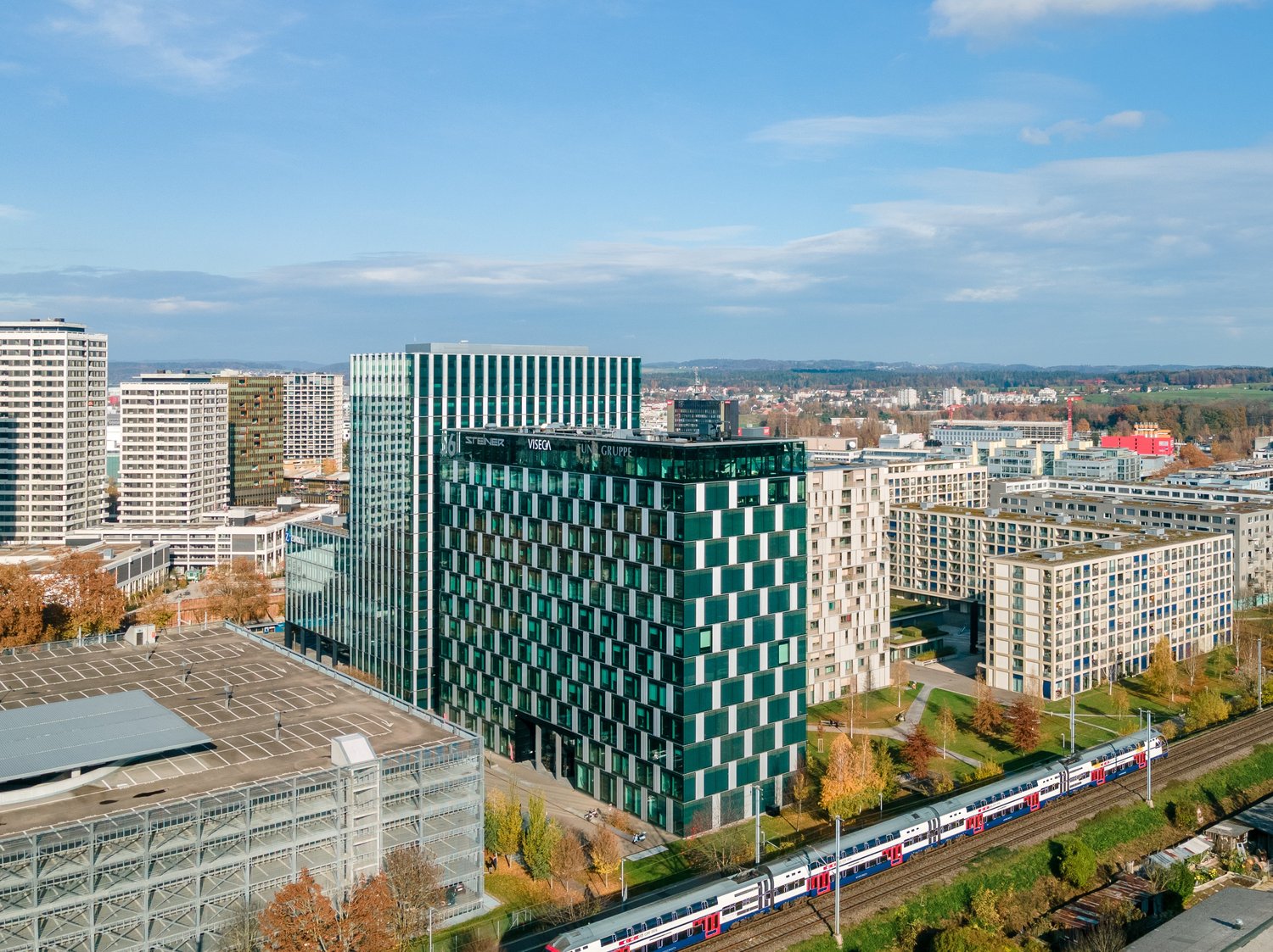 The image shows a cityscape with a mix of high-rise buildings, including a large green-and-white office building in the center. There are also several other tall buildings and a train passing through the scene. The surrounding area appears to be a mix of