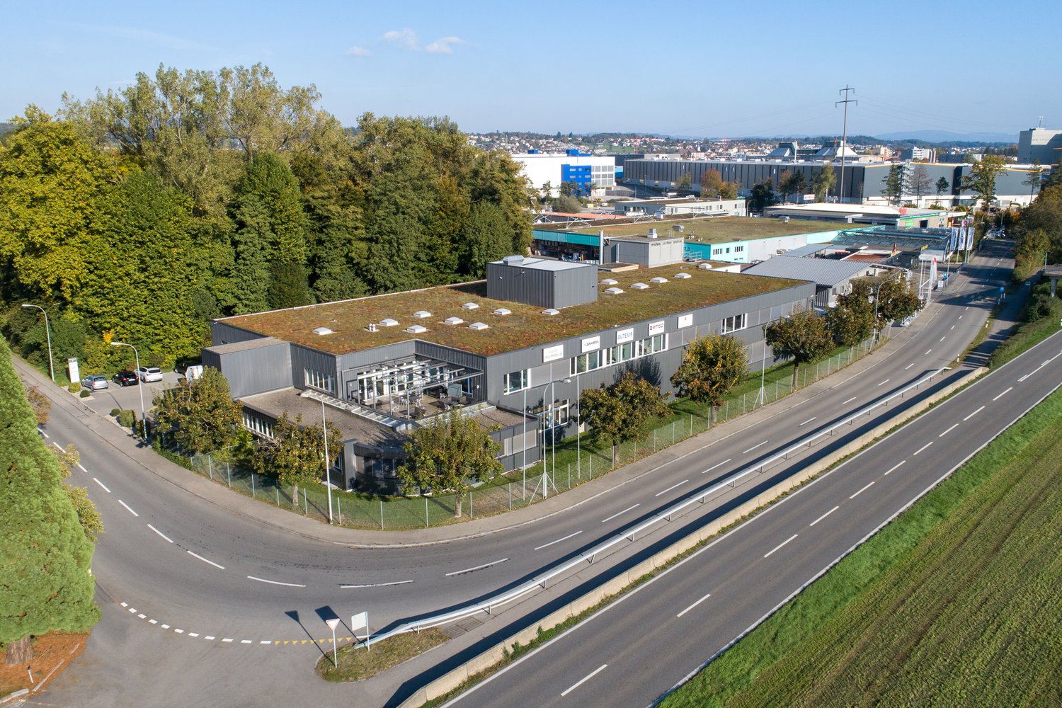 Large commercial building with a green roof, surrounded by trees and a road with multiple lanes. The building appears to be an industrial or office complex with parking spaces visible in the foreground.