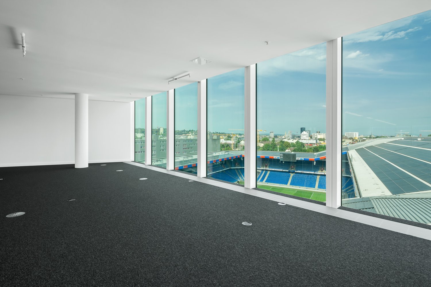 Large open office space with floor-to-ceiling windows overlooking a sports stadium and city skyline. The space has a black tile floor and white walls. There are several lighting fixtures installed in the ceiling.